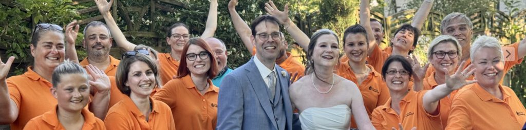 Herzergreifende Freude: Der JungerChor TakeFour posiert fröhlich in orangen Shirts mit dem strahlenden Brautpaar im Garten des Weinguts – alle lachen und winken begeistert in die Kamera.