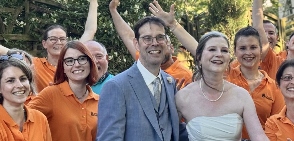 Herzergreifende Freude: Der JungerChor TakeFour posiert fröhlich in orangen Shirts mit dem strahlenden Brautpaar im Garten des Weinguts – alle lachen und winken begeistert in die Kamera.