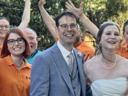 Herzergreifende Freude: Der JungerChor TakeFour posiert fröhlich in orangen Shirts mit dem strahlenden Brautpaar im Garten des Weinguts – alle lachen und winken begeistert in die Kamera.