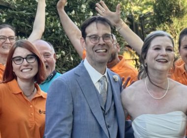 Herzergreifende Freude: Der JungerChor TakeFour posiert fröhlich in orangen Shirts mit dem strahlenden Brautpaar im Garten des Weinguts – alle lachen und winken begeistert in die Kamera.