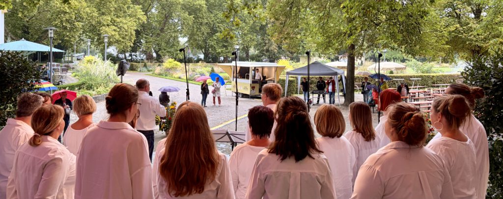 Trotz Regen: TakeFour-Chorkonzert beim Oktoberfest im Ebertpark Ludwigshafen   Chor TakeFour LudwigshafenBlick von der Bühne: Der JungerChor TakeFour singt beim Oktoberfest 2025 im Ebertpark Ludwigshafen. Die Chormitglieder stehen in weißen Hemden mit dem Rücken zur Kamera, vor der Bühne sitzen und stehen Zuhörerinnen und Zuhörer teils unter Regenschirmen. Im Hintergrund sind Verkaufsstände und Bäume im Park zu sehen.