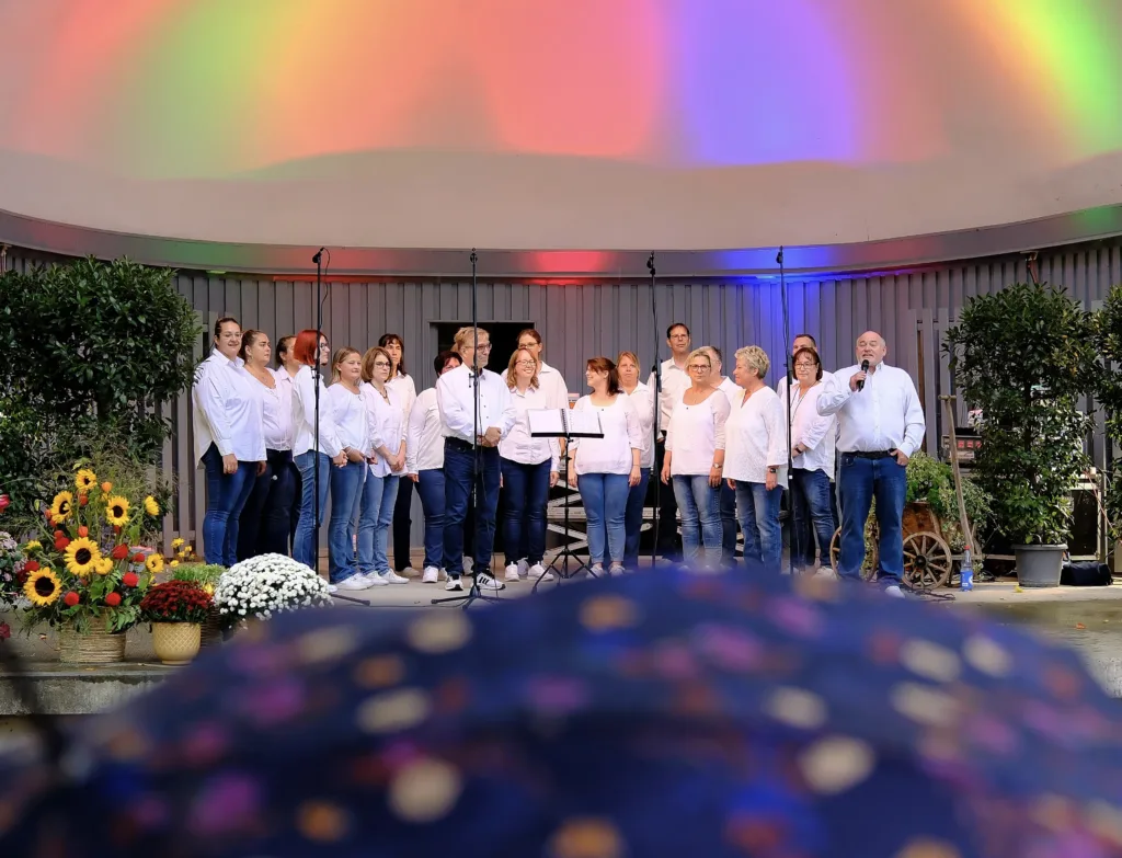 Der JungerChor TakeFour singt beim Oktoberfest 2025 in der Konzertmuschel im Ebertpark Ludwigshafen. Die Chormitglieder stehen in weißen Hemden und Jeans auf der Bühne, Chorleiter Klaus Hoffmann spricht ins Mikrofon. Im Vordergrund sind Regenschirme der Zuschauer zu sehen, bunte Lichtprojektionen färben die Muschel in Regenbogenfarben