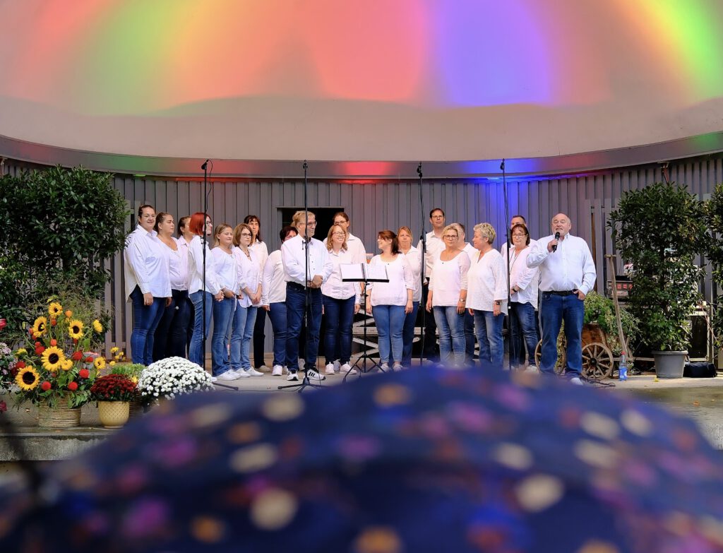 Der JungerChor TakeFour singt beim Oktoberfest 2025 in der Konzertmuschel im Ebertpark Ludwigshafen. Die Chormitglieder stehen in weißen Hemden und Jeans auf der Bühne, Chorleiter Klaus Hoffmann spricht ins Mikrofon. Im Vordergrund sind Regenschirme der Zuschauer zu sehen, bunte Lichtprojektionen färben die Muschel in Regenbogenfarben